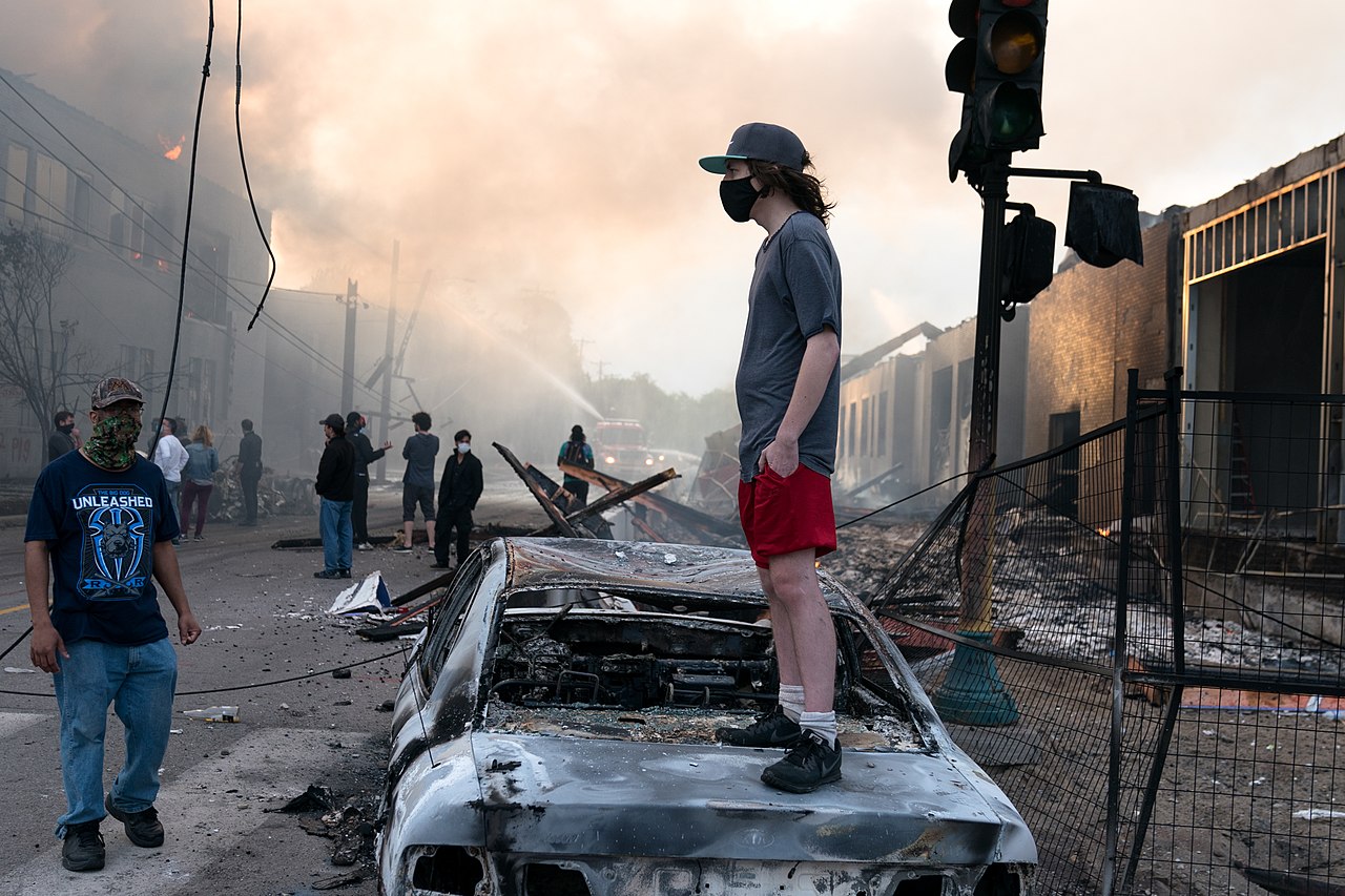 1280px-A_man_stands_on_a_burned_out_car_on_Thursday_morning_as_fires_burn_behind_him_in_the_Lake_St_area_of_Minneapolis,_Minnesota_(49945886467)